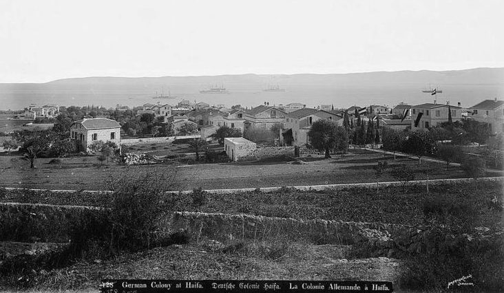 Black and white photograph seemingly shred off at the top. Across the center is a large grouping of two- to three-story buildings with slightly sloped roofs, surrounded by scattered trees and grass or earthen lots. The buildings sprawl out to the background, down against a sea wherein there are four boats. In the foreground of the image is a grassy field with bushes and small trees and two stone walls splicing across it.