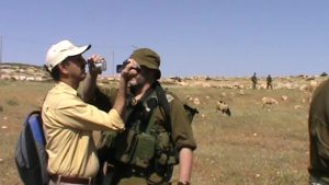 In a mild desert landscape, a man in a yellow shirt and white baseball cap and holding a blue backpack points a camera in the face of another man. The other man is dressed in army greens and points a camera in the face of the man in the yellow. 