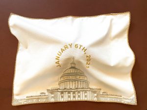 A brocade challah cover with gold trim is on a reddish brown background. The challah cover is brightly lit and is propped up by the challah that it covers underneath. The challah is not visible. The fabric is white and the US Capitol is embroidered in gold in the center. Above the Capitol, “JANUARY 6TH, 2021” is embroidered in gold in a circular shape above and around the Statue of Freedom. Below the dome, the hebrew phrase חדש ימינו כקדם is embroidered in gold.