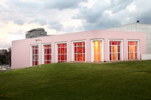 The exterior of the gallery viewed during the day. Sunlight penetrates into the gallery through the windows and the interior is visibly red from the outside.