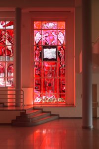 The first window depicts a sort of an expulsion scene painted in deep red. At the center of the window is an open book with Hebrew text written on both sides (quoted from ‘Ro’achem,’ a book the artist wrote about seven years earlier). On the right side, the text is written as normal, from right to left, but on the left side, it is written in mirror to the other side, from left to right. Next to the image of the book, on both sides of it, appear two semi-human figures, both pointing at it with their hands. Their feet look like the legs of a beast. Above the image of the book, two angels appear, facing each other, also gesturing toward the book below. Above the angels, at the top of the painting, there is a big eye with curved rays coming out of it and a light bulb within — a reference to Picasso’s “Guernica.” Underneath the book and coming down from it is a ladder that gets more and more narrow as it goes down to the bottom of the window. On the ladder is a figure of a naked and hairy man climbing downward. At the bottom of the window there is a winding tangle of tails and scales, headless and lacking any other organs.