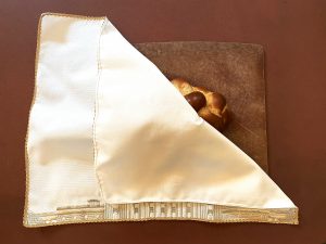 A brocade challah cover with gold trim is on a reddish brown background. The challah cover is brightly lit and the upper right hand corner of the cover is folded open revealing a braided challah underneath. The challah is on a brown cutting board with numerous knife marks. At the bottom of the challah cover, a small section of the Capitol Building is visible under the folded fabric. 