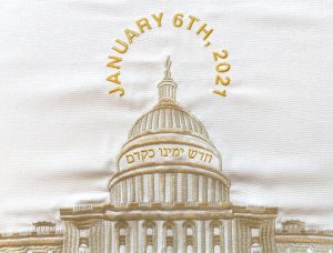 A detail photograph of a brocade challah cover. The fabric is white and the US Capitol is embroidered in gold in the center. Above the Capitol, “JANUARY 6TH, 2021” is embroidered in gold in a circular shape above and around the Statue of Freedom. Below the dome, the hebrew phrase חדש ימינו כקדם is embroidered in gold.
