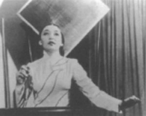 Blurred black-and-white photograph of Clara Rockmore playing the theremin. She stands before the black, waist-high electronic instrument as if before a keyboard, long dark hair stroked tight back behind her head, dark eyes and lips pointed up, wearing a light full-sleeved gown. Her right hand is lifted up before her right shoulder, gripping a small black electronic device, while her left hand hovers out beyond the left end of the theremin. The shadows of electronic cables or the cables themselves cross her upper body, perhaps connected to the device in her right hand.