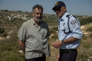 A middle-aged man in plain clothes, Hussein, is on the left side of the image while Avi Levi, a police official in uniform, discusses with him to the right. A landscape of Jerusalem hills fills the background.