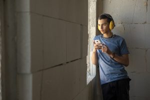 A young teenager, Muhammad, leans against a cement wall while holding a cell phone. He looks out a window in the wall, but what he is looking at is not visible. The walls look unfinished and Muhammad wears yellow construction earmuffs.