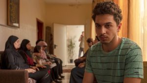 A young man, maybe late teens or early 20s with curly hair and a very light moustache and beard appears to be watching something, maybe a TV, that is not shown. Behind him sit about 6 women in hijab on a couch and looking down or with their eyes closed.