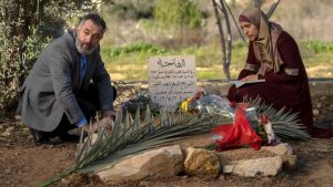 A middle-aged man, Hussein, crouches down at the left side of a grave. He wears a gray suit with a bright blue shirt. A woman crouches on the opposite side of the grave — Suha, Muhammad’s mother. She wears a scarlet red thobe and a beige hijab. There are flowers around the grave and Suha rests a book in her crouched lap. 