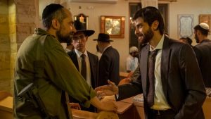 The setting appears to be a synagogue. On the left, a man, Simon, is wearing a kippah and an army uniform with a gun hanging off his arm. He’s talking to another man, Yosef ben Haim, who is wearing a suit, tie, and kippah and who holds out his hand towards Simon to shake it. A younger man, Avishai, in a black hat looks on. In the background, there are a number of men in the synagogue, diversely dressed; some wear black hats and others wear white kippot.