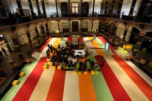 The camera looks down on a large, inner court of an exhibition hall. At the center is a table surrounded by scholars. The flooring is made of thick strips of green, red, white, and orange. An arched hallway lines the court and is lit from within.