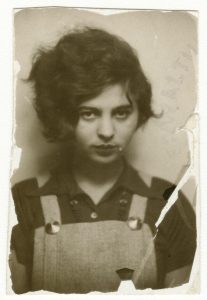 Old, rectangular and sepia-colored photograph of a young Mascha Kaléko staring directly at the viewer. The edges of the photograph are torn or rotted off, while a tear cuts across the bottom-right corner, down through her shoulder. Kaléko has very thick, wavy dark hair, the whole of which is combed entirely to the right side of her head, framing the side of her face. She wears grey, perhaps wool overalls, and a darker colored short-sleeved, collared shirt beneath. Her gaze is soft and firm at the same time.