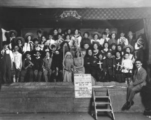 A black and white cast photo shows Purim play actors dressed in costume and assembled on stage, facing the camera. On the right, the director sits on the stage ledge. A sign in Yiddish hangs center next to a short ladder providing steps from the ground to the stage platform.
