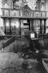 Black and white interior view of the Talmud Torah Synagogue in partial ruin. The ark is center, opened and empty. Dirt covers the floor.