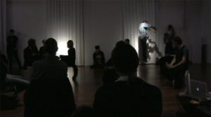 In a dimly lit room, a figure seems to levitate. Propelled vertically into the air, his arms are outstretched from the shoulder. An audience of people at a variety of vantage points, seated on the floor, on chairs, and standing, all turn to focus on the floating figure. 