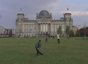 Under a lavender and pale blue dusky sky, on a vast green front lawn, opposite the Reichstag. Visible in the distance on either side are a few decorative trees with autumn leaves in gold, red and green. In the background, friends kick a soccer ball, people stroll through the grounds. In the foreground a figure in a blue and green plaid flannel and black trousers while pressing down on his knee curves his entire body sideways into an unexpected half circle shape.