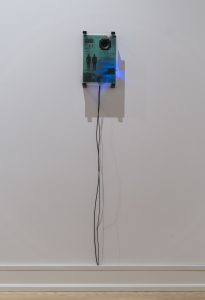 In the center of the image is one green satin acrylic board with exposed wiring on a white wall. Black wires extend from the bottom of the board and are tucked into the white floorboard. There is a faint blue light emanating from behind the satin acrylic board.