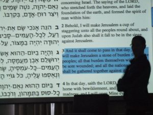 A figure in silhouette turns their head sharply towards the camera and away from the screen projection in front of which they stand with arms crossed over their chest. The projection is a page of parallel bilingual text, English on one side Hebrew on the other, and one verse is highlighted. The highlighted verse is the prooftext in the book of Zecharia from which the halacha of mechitza is derived.