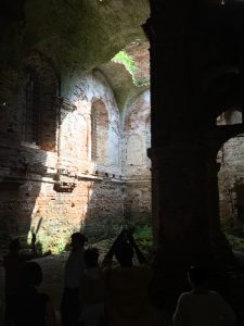 Inside the ruins of a synagogue. The brick walls are intact but light pours in from an unseen hole in the roof. Moss grows on the walls. At the bottom of the photograph, in shadows, are human figures. They are holding their hands up as if they are taking a photo of the light with their phones.