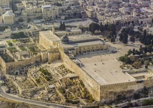 An aerial view of Al-Aqsa Mosque on the Temple Mount in the Old City of Jerusalem. The compound, built with Jerusalem limestone, is bathed in bright sunlight and fills the majority of the image. City buildings are in the background at the top of the image. A road is in the lower left corner and continues along the center at the bottom of the image. 