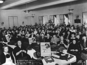 Black and white photograph of a mass of women knitting. They are seated in rows, facing the camera. A table in front of the rows of women knitting, and behind two women in the foreground, displays posters soliciting donations for "solidary gifts" to soldiers fighting the Nazis. Text written onto the photograph along the bottom reads, "International Workers Order Knitting Circle Luncheon, Los Angeles, Nov. 12, 1941.