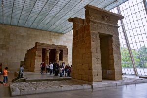 Two ancient-looking, stone structures are in a room at the Met Museum with a floor-to-ceiling glass window. One of them has two columns in front. The other has a rectangular opening. A group of people stand in between them. 