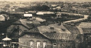 A photographic view of the rooftops of Bobruisk. In the foreground is a large, wooden structure. In the background is a road that winds to the top-right corner before turning and following the top horizontal border of the postcard.