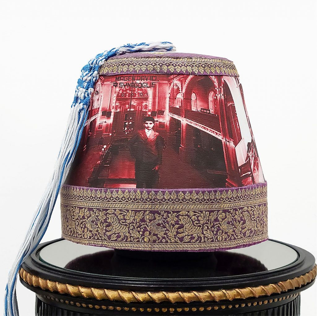 A burgundy fez cap with an Indian silk border embroidered in gold is perched on top of a round black pedestal. Pictured on the face of the hat is a solemn young boy confidently standing in the sanctuary of a Sephardic synagogue, looking out at the viewer. At the upper left is a young woman standing on the second floor gallery. The customary tassel at the top of the fez cap is replaced by blue and white tzitzit (ritual fringes or tassels) knotted in the Sephardic style.